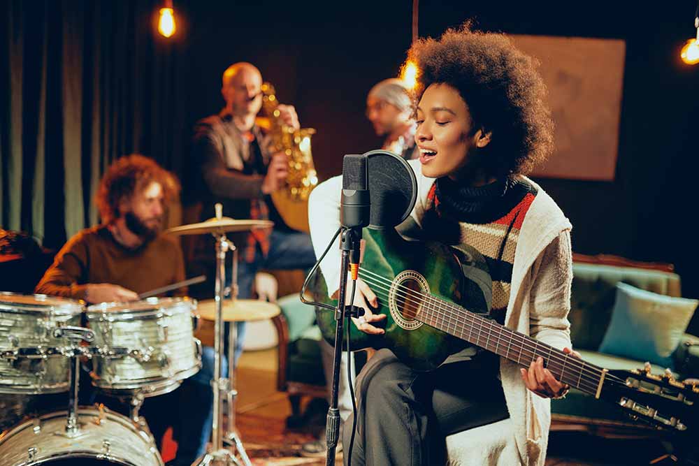 Woman singing and playing a guitar while sitting on chair in a recording studio, along with a drummer, saxophonist and bass guitarist.