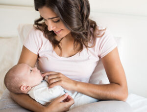 A smiling mother in a pink shirt cradling her newborn baby on a nursing pillow during a breastfeeding support group class.