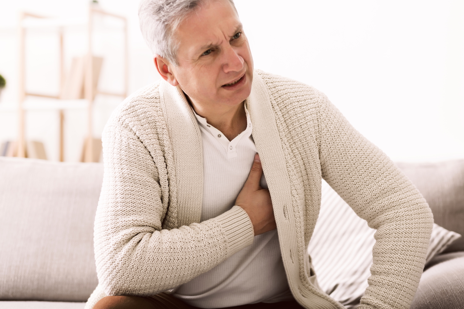 Senior man with chest pain sits on couch with his hand on his chest.